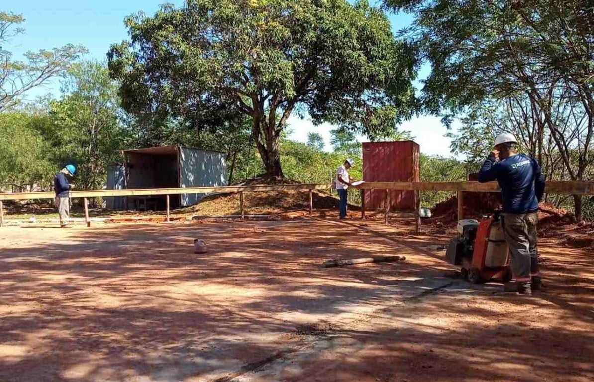 Obras da Cidade das Crianças em Jundiaí, mostrando trabalhadores em um canteiro de obras cercado por árvores, com contêineres ao fundo.