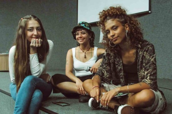 Três jovens sorrindo sentadas no palco durante o evento "Juventudes na Correria" no Sesc Jundiaí, refletindo diversidade e descontração.