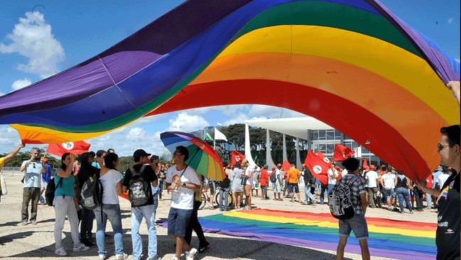 Grupo de pessoas segurando uma grande bandeira de arco-íris, que é símbolo do movimento LGBTQIA+.