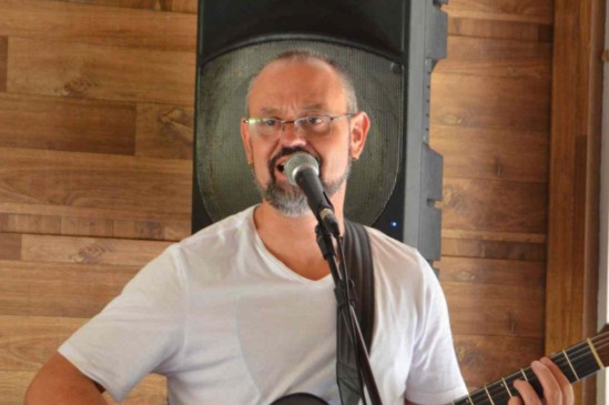 Cantor Tom Nando se apresenta com violão e microfone, usando camiseta branca, em ambiente com painel de madeira ao fundo.