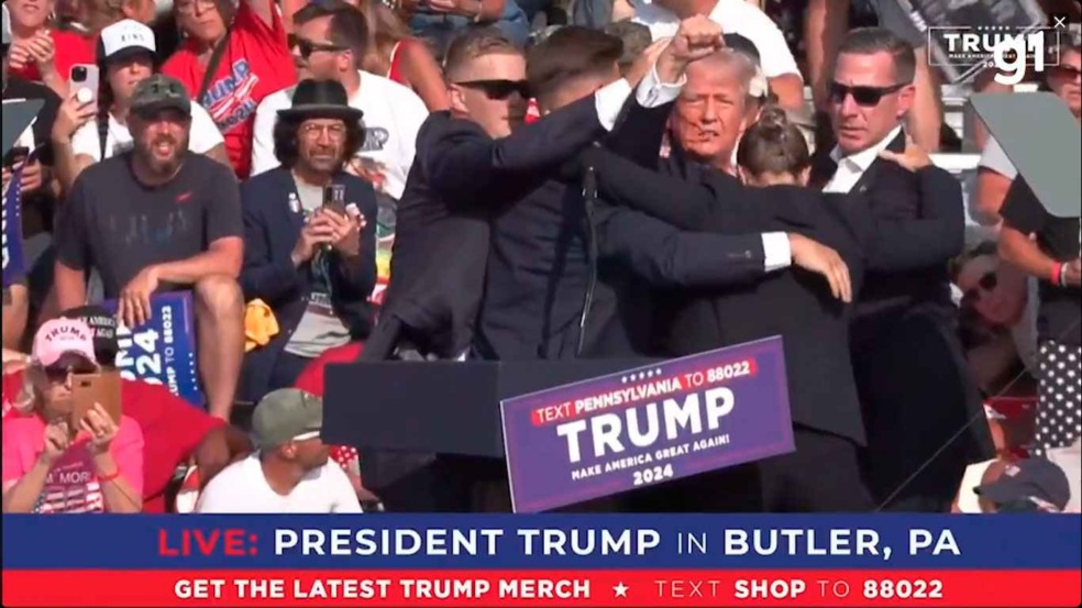 A imagem mostra Donald Trump sendo escoltado por seguranças durante um comício em Butler, Pensilvânia, após um atentado.