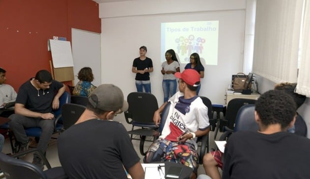 Foto de sala com jovens, três deles estão à frente apresentando trabalho