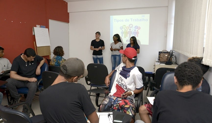 Foto de sala com jovens, três deles estão à frente apresentando trabalho