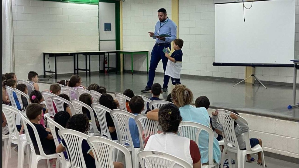 Um homem com um menino na frente de uma plateia de crianças, possivelmente em um evento de educação financeira.