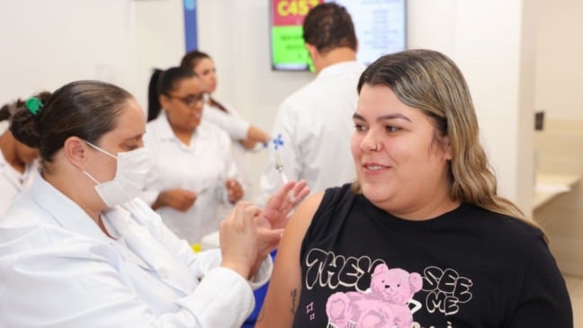 Uma mulher recebe uma injeção no braço enquanto outra mulher em uniforme médico segura a seringa.