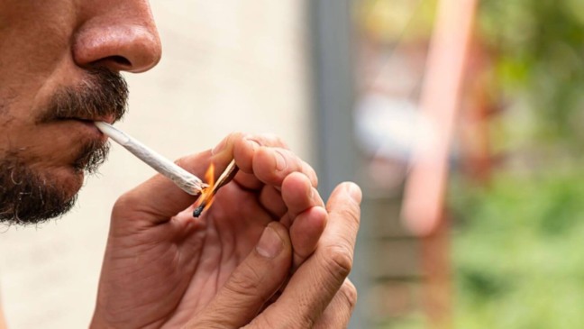 Homem acendendo um baseado de maconha ao ar livre, close-up no rosto e nas mãos, destacando o uso da substância.