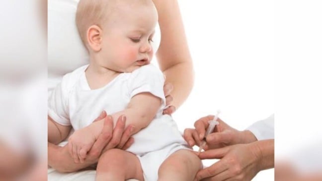 Segurado pela mãe, bebê observa enquanto mãos de enfermeira aplicam vacina em sua perna esquerda