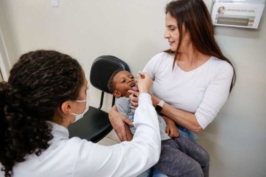 Mãe segurando criança enquanto profissional de saúde aplica Vacina contra a Paralisia Infantil em UBS