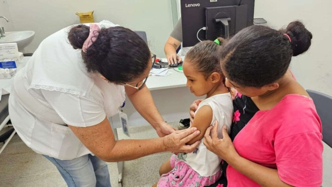 Enfermeira aplicando dose de vacina em menina, que está sentada no colo de responsável.
