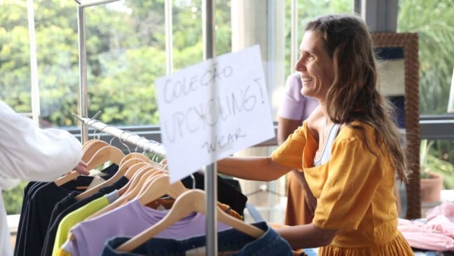 Mulher de vestido amarelo sorri ao organizar roupas coloridas em uma arara com placa escrita “Coleção Upcycling Wear”, em feira de moda sustentável no Sesc Jundiaí.