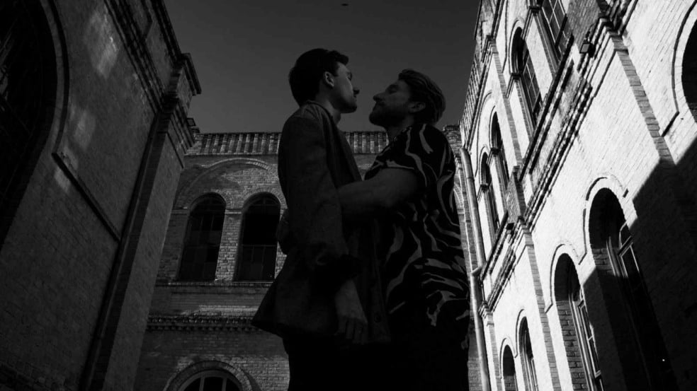 Dois homens se encaram de perto em ambiente urbano com prédios de tijolos, em fotografia preto e branco de estilo dramático.