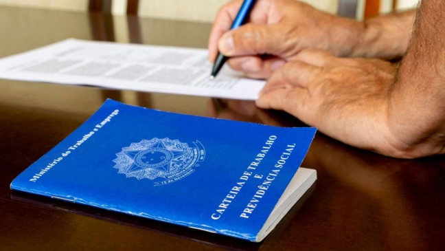Carteira de Trabalho brasileira em destaque sobre a mesa, enquanto um trabalhador assina contrato ao fundo.