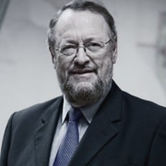 Professor Antônio Carbonari Neto, de terno escuro e gravata azul, sorri diante de fundo claro.