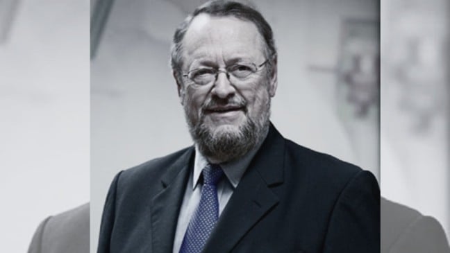 Professor Antônio Carbonari Neto, de terno escuro e gravata azul, sorri diante de fundo claro.