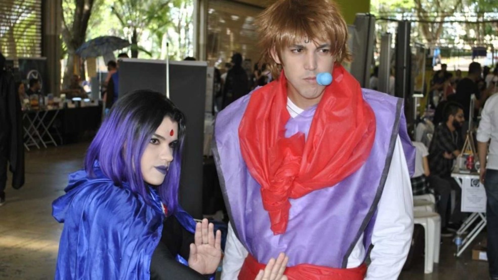 Duas pessoas em evento de cultura pop fazendo cosplay: à esquerda, personagem de capa azul e cabelo roxo; à direita, personagem com roupas lilás e vermelhas, peruca castanha e bola azul na boca. Ao fundo, público e estandes do evento. Duas pessoas em evento de cultura pop fazendo cosplay: à esquerda, personagem de capa azul e cabelo roxo; à direita, personagem com roupas lilás e vermelhas, peruca castanha e bola azul na boca. Ao fundo, público e estandes do evento.