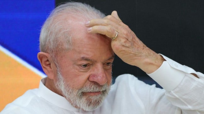 Presidente Lula com expressão séria e a mão na cabeça durante evento, usando camisa branca.