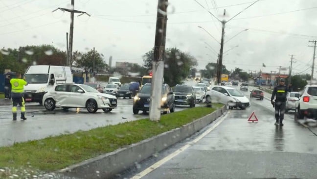 Agentes de trânsito controlam o fluxo de veículos em uma via movimentada sob chuva, após um acidente envolvendo dois carros brancos; um deles está atravessado na pista e o outro sobre o canteiro central.