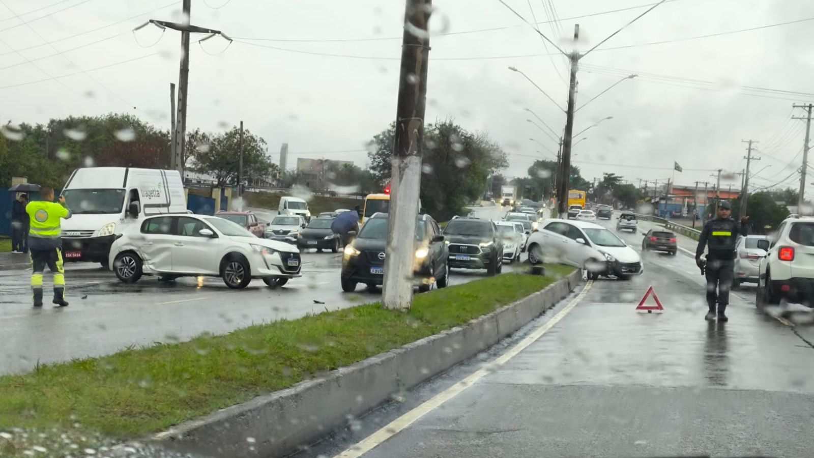 Agentes de trânsito controlam o fluxo de veículos em uma via movimentada sob chuva, após um acidente envolvendo dois carros brancos; um deles está atravessado na pista e o outro sobre o canteiro central.