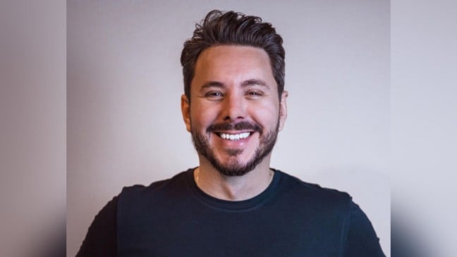 Alan Barros sorrindo, de barba curta e cabelos escuros penteados, vestindo camiseta preta diante de fundo claro.