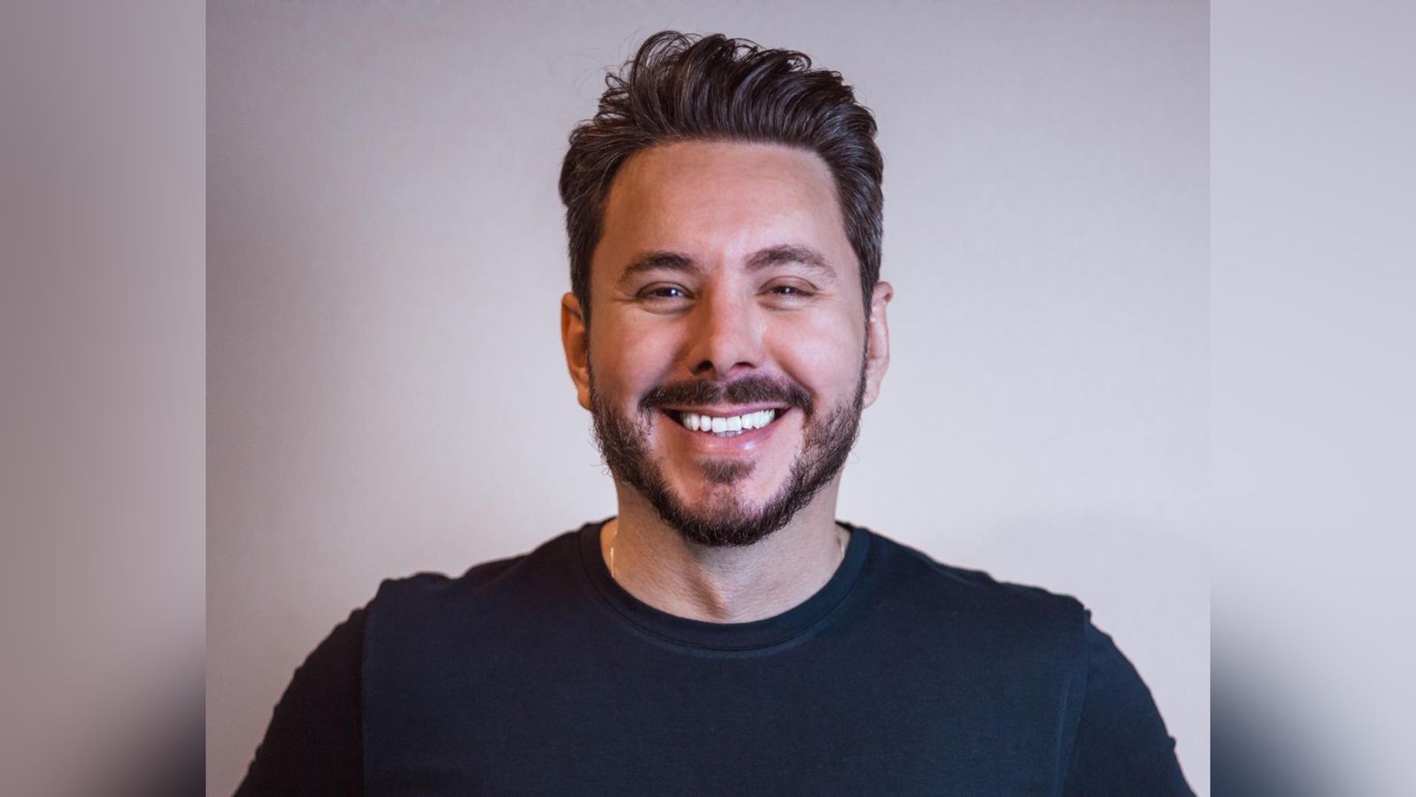 Alan Barros sorrindo, de barba curta e cabelos escuros penteados, vestindo camiseta preta diante de fundo claro.