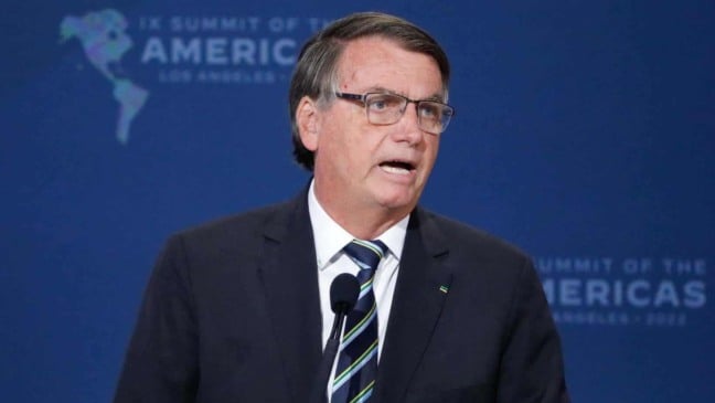 Ex-presidente Jair Bolsonaro falando em púlpito, usando terno escuro e gravata listrada, diante de fundo azul com o logotipo da Cúpula das Américas.