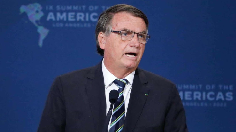 Ex-presidente Jair Bolsonaro falando em púlpito, usando terno escuro e gravata listrada, diante de fundo azul com o logotipo da Cúpula das Américas. Ex-presidente Jair Bolsonaro falando em púlpito, usando terno escuro e gravata listrada, diante de fundo azul com o logotipo da Cúpula das Américas.