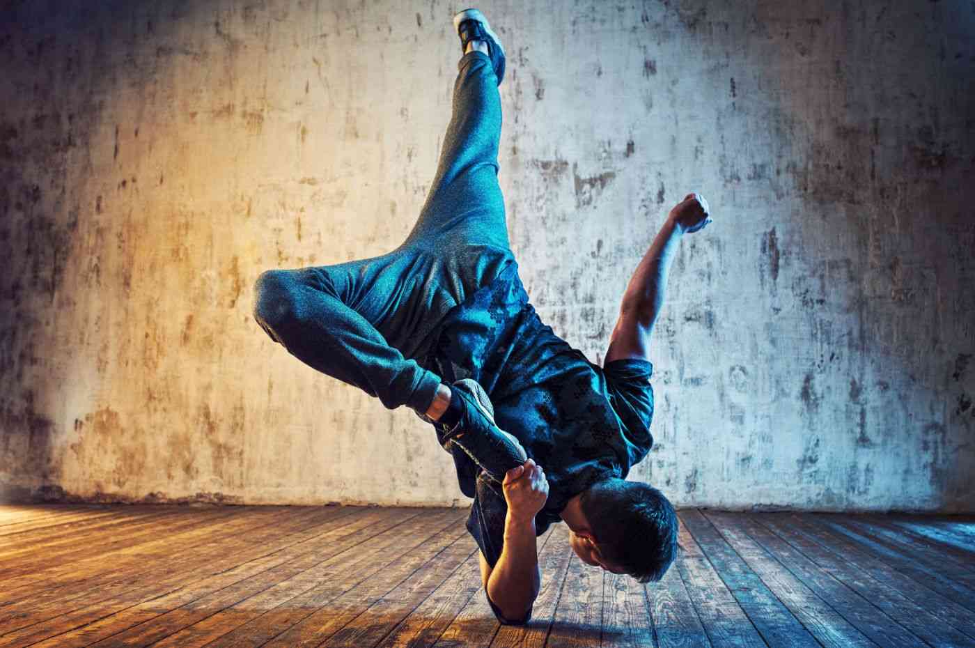 Dançarino de breaking executa movimento acrobático com uma mão no chão e pernas no ar em estúdio de dança com fundo industrial.
