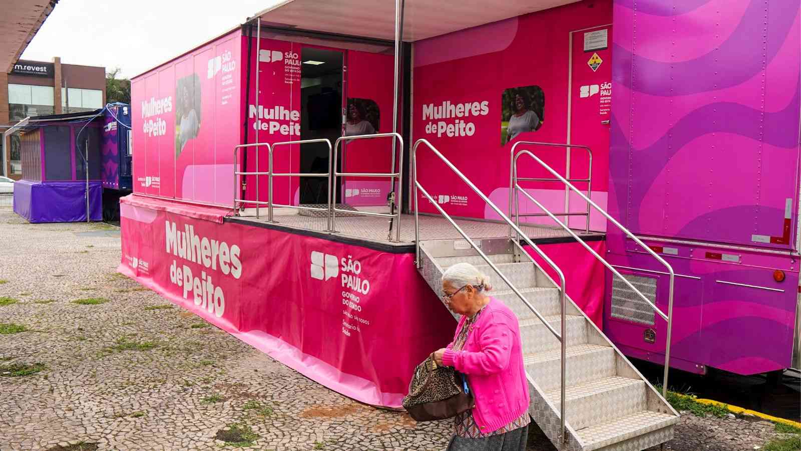 Carreta da Mamografia do programa Mulheres de Peito, em Várzea Paulista, com estrutura rosa e rampa de acesso, enquanto uma mulher idosa passa em frente.