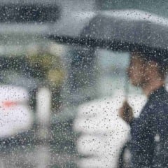 Pessoa caminha com guarda-chuva sob chuva em Jundiaí, vista através de janela com gotas de água.