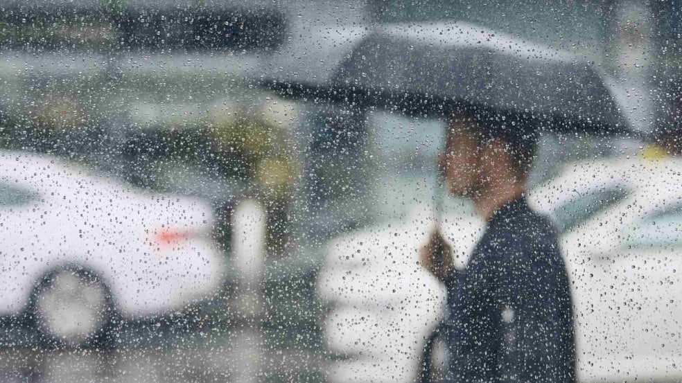 Pessoa caminha com guarda-chuva sob chuva em Jundiaí, vista através de janela com gotas de água.