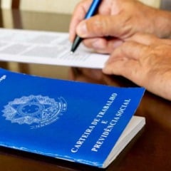 Carteira de Trabalho azul ao lado de pessoa assinando contrato de emprego sobre mesa de madeira.
