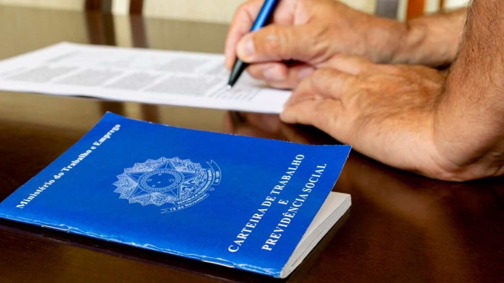 Carteira de Trabalho azul ao lado de pessoa assinando contrato de emprego sobre mesa de madeira.