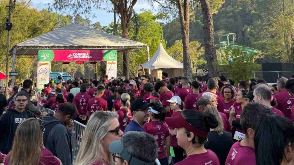 Atletas concentrados na largada da 1ª Corrida das Orquídeas em Várzea Paulista, no Clube de Campo da Cica, com participantes vestindo camisetas bordô e arco de largada ao fundo.