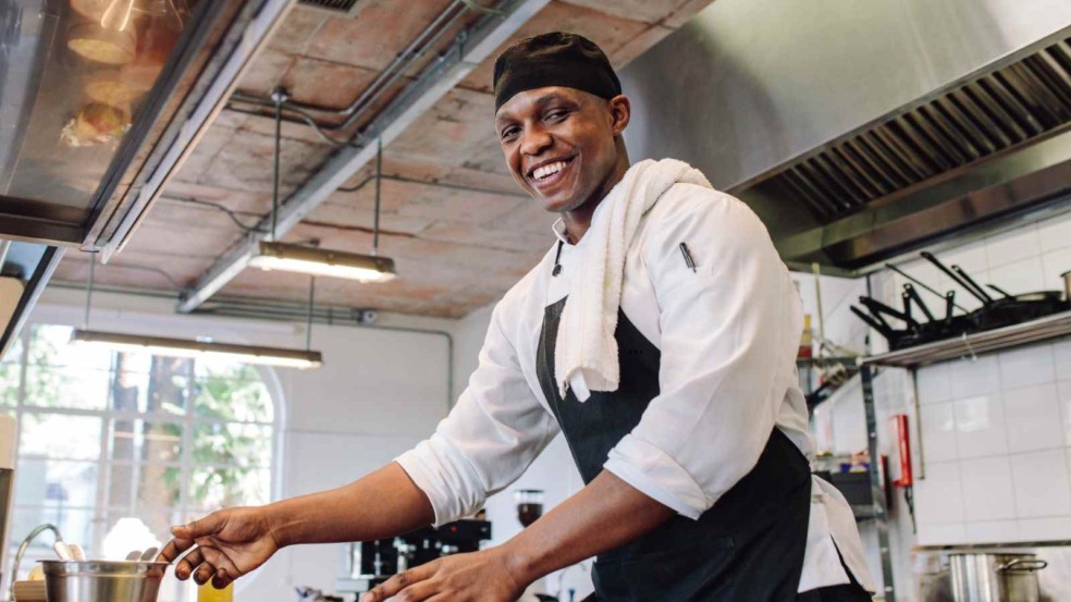 Homem negro sorridente com uniforme de cozinha e avental preto mexendo uma panela em cozinha industrial. Homem negro sorridente com uniforme de cozinha e avental preto mexendo uma panela em cozinha industrial.