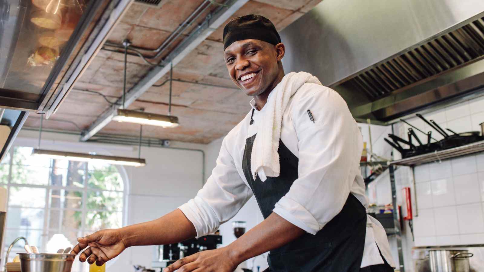 Homem negro sorridente com uniforme de cozinha e avental preto mexendo uma panela em cozinha industrial.