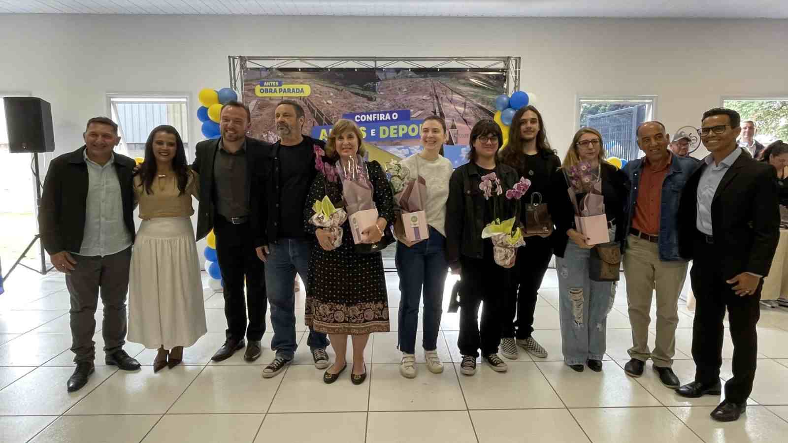 Autoridades e familiares posam para foto durante a inauguração do CEMEB Professora Adriana Tozetto em Itupeva, com flores e painel “Antes e Depois” ao fundo.
