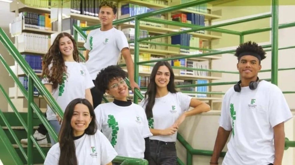 Estudantes do IFSP posando em escada no campus, com camisetas do Instituto Federal e biblioteca ao fundo.
