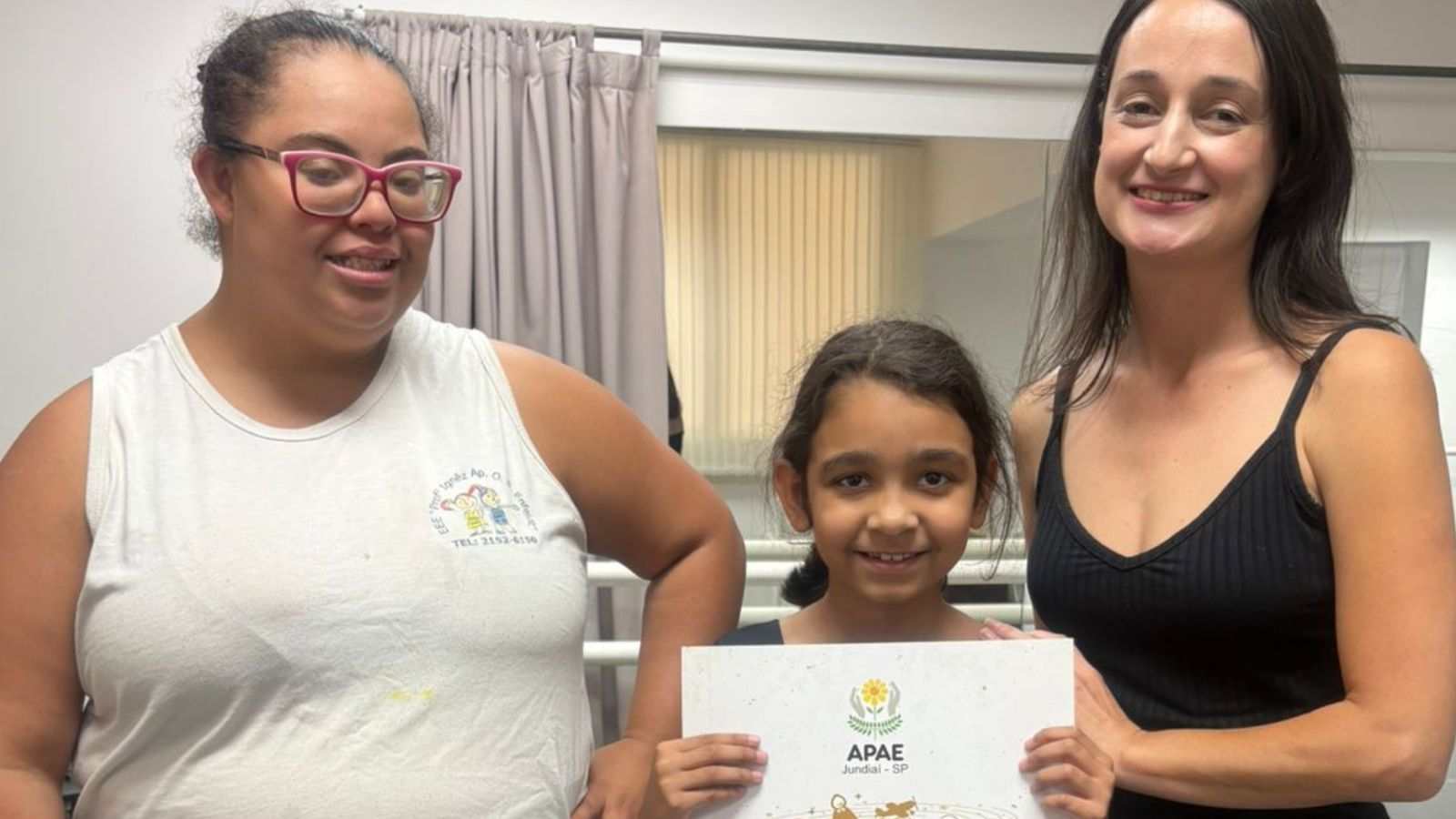 Professora de dança da APAE de Jundiaí, Raquel Tumolo Leopoldino, e duas alunas segurando um certificado da instituição e sorrindo durante ensaio do espetáculo “Dentro da História”.