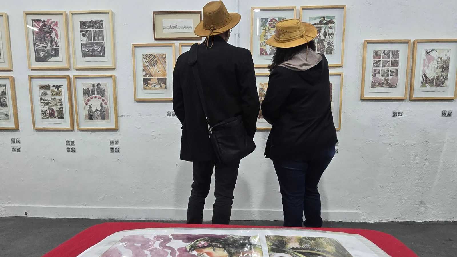 Dois visitantes de chapéu observam páginas originais emolduradas da HQ A Viola Encarnada, expostas na mostra Nos Braços do Violeiro em Bragança Paulista.