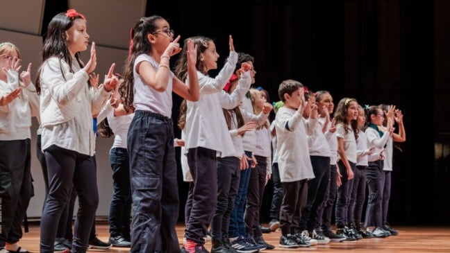 Crianças em fila no palco durante apresentação coral, usando roupas em branco e preto e gesticulando com as mãos em sincronia.