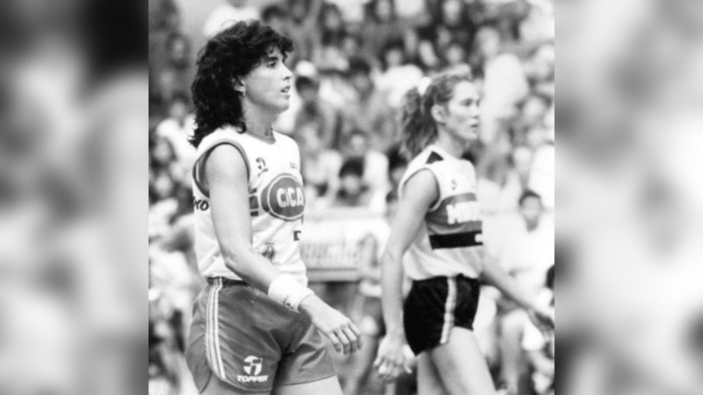 Foto em preto e branco de duas jogadoras de vôlei em quadra; à frente, atleta com uniforme da Cica, e ao fundo, outra jogadora observando o jogo.