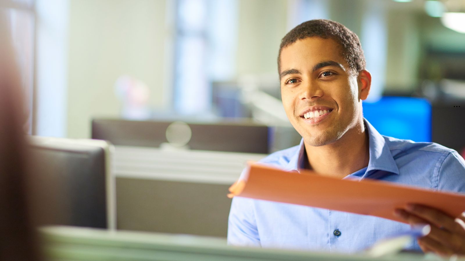 Jovem estagiário sorridente em escritório, segurando pasta laranja durante atendimento.