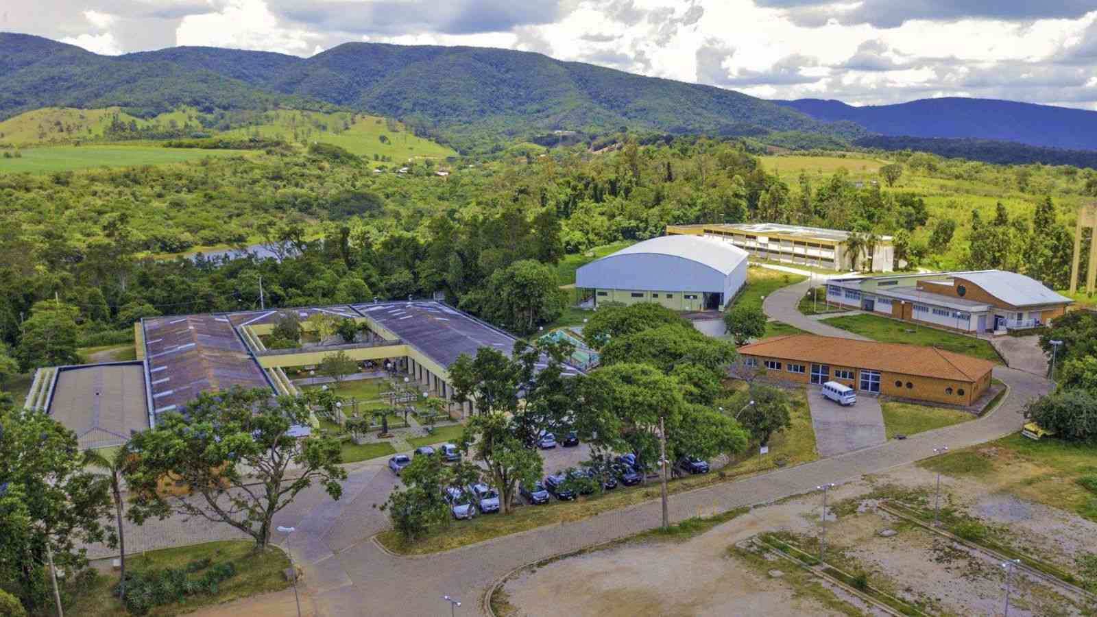 Vista aérea do campus da Etec Benedito Storani, em Jundiaí, cercado por áreas verdes e montanhas ao fundo. A imagem mostra os prédios da escola, ginásio, estacionamentos e áreas de convivência, destacando a integração da instituição com o ambiente natural.