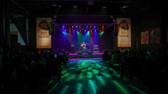 Artista toca violão no palco iluminado do Festival de Música de Jundiaí, com público assistindo na Sala Glória Rocha.