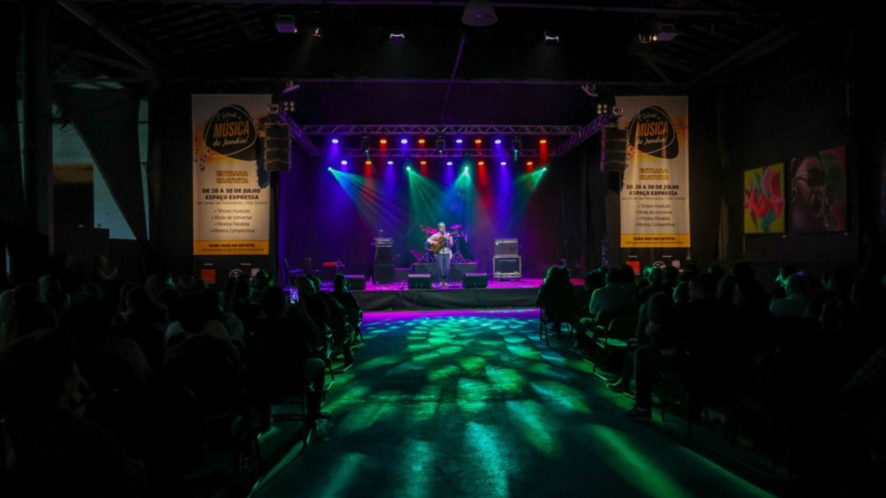 Artista toca violão no palco iluminado do Festival de Música de Jundiaí, com público assistindo na Sala Glória Rocha.
