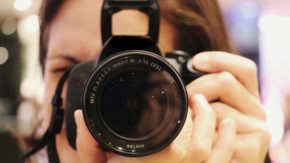 Close de uma pessoa segurando uma câmera fotográfica, com foco na lente em primeiro plano. Close de uma pessoa segurando uma câmera fotográfica, com foco na lente em primeiro plano.