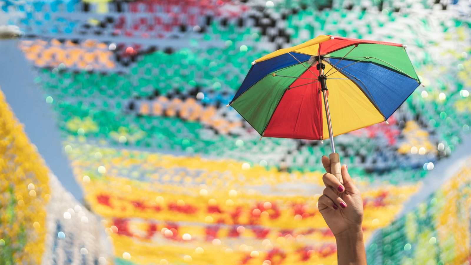 Mão segurando um guarda-chuva colorido de frevo com fundo de bandeirolas coloridas em clima de festa popular.