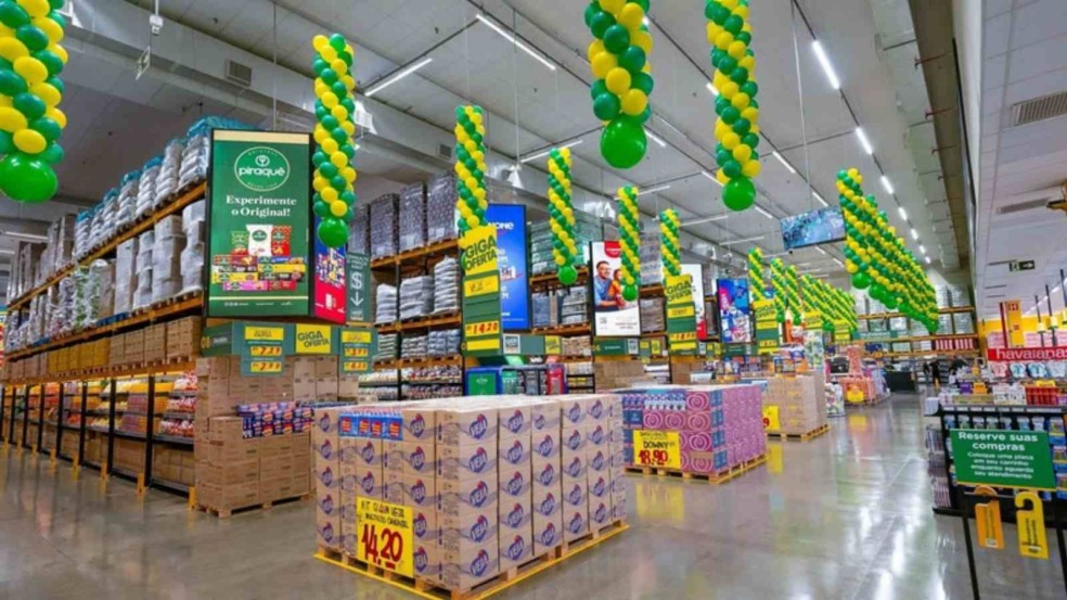 Interior da loja Giga Atacado decorada com balões verdes e amarelos, prateleiras cheias de produtos e promoções em destaque nos corredores. Interior da loja Giga Atacado decorada com balões verdes e amarelos, prateleiras cheias de produtos e promoções em destaque nos corredores.