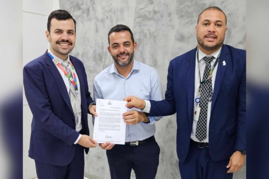 Gustavo Martinelli ao centro segura um documento oficial entre dois outros homens de terno, representando reunião formal para apresentação de ofício.