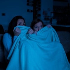 Duas mulheres com olhar assustado assistem a um filme de terror cobertas por um cobertor azul.